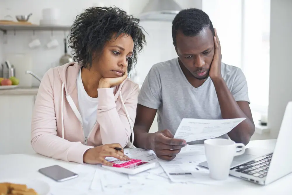 Stressed african american couple looking frustrated, having no money to pay off their debts, managing family budget together, sitting home at kitchen table with lots of papers, laptop and calculator